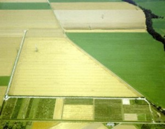 Luftbild mit Feldern, Weiden und Wiesen; entlang eines Feldweges laufen zwei lange weiße Röhren rechtwinklig aufeinander zu