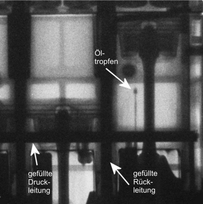 Einzelbild aus dem Film des untertourig bewegten Motors Schwarz-Weiß-"Foto" (Neutronenradiographie) eines Motorausschnitts. Man erkennt zwei Kolben, die auf unterschiedlicher Höhe stehen, einen Ölstrahl, der von unten nach oben schießt, und senkrecht und waagerecht verlaufende schwarze Balken, die die mit Öl gefüllten Druck- und Rückleitungen sind.