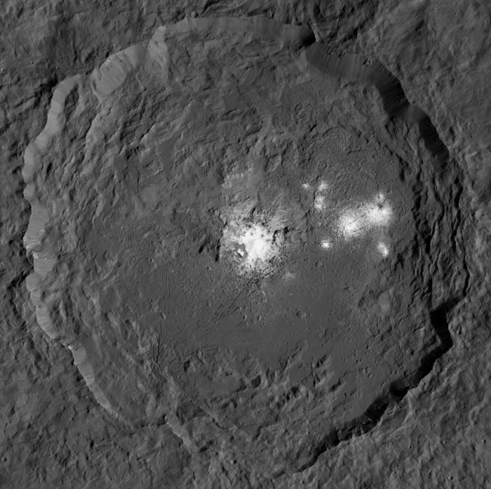 Eine dunkle Planetenoberfläche, im Bild ein tiefer Krater mit weich auslaufenden Rändern. Im Zentrum des Kraters eine helle, von Furchen durchzogene, runde Region, die am Rand unregelmäßig ins Gestein ausläuft.