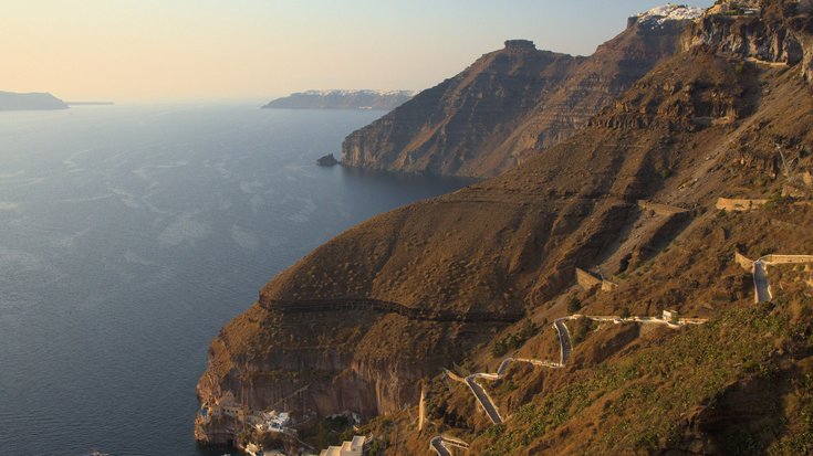 Links das Mittelmeehr und rechts die ziemlich steile Caldera-Wand. Unten am Meer sind weiße Gebäude. Ein Pfad führt den Berg hoch, wo ebenfalls weiße Gebäude stehen. Im Hintergrund sind noch mehr Teile der Inselgruppe zu sehen