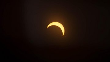 Fotoaufnahme einer partiellen Sonnenfinsternis: Die leuchtende Sonnenscheibe ist links oben von einem dunklen, kreisrunden Schatten des Mondes verdeckt, sodass eine sichelförmige, leuchtende Fläche sichtbar bleibt.