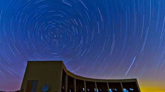 Gebäude mit vielen Teleskopen, darüber der Nachthimmel mit Sternspuren.