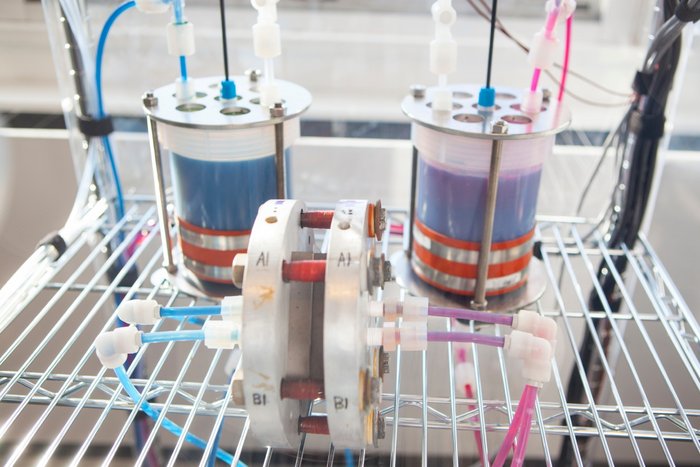 Flussbatterie als Windstromspeicher Verkabelter Aufbau einer Batterie mit zwei zylinderförmigen Gefäßen im Hintergrund und zwei zusammenhängenden Metallscheiben im Vordergrund.