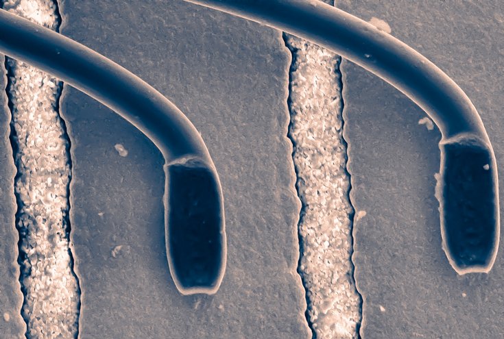 Mehrere gebogene, röhrenförmige Leiterbahnen verlaufen in Kurven über eine körnige Oberfläche. Die hakenförmigen Strukturen zeigen die filigrane Mikroarchitektur des Chips.