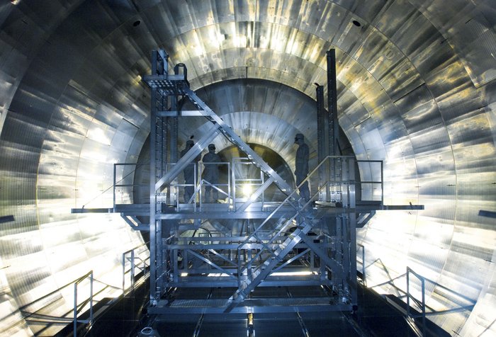 Das Spektrometer wird installiert Das Spektrometer am KIT ist 24 Meter lang und hat einen Durchmesser von 10 Metern. Die Aufnahme zeigt eine Frontalansicht. Auf dem Gerüst davor befinden sich drei Menschen in Schutzkleidung.
