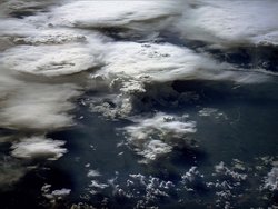 Foto. Blick aus dem Weltall auf die Erdoberfläche. Man erkennt zahlreiche Wolken, die sich zum Teil als weiße kreisförmige Flächen ausdehnen.