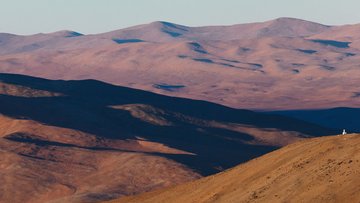 Vor dem Hintergrund einer bergigen Wüstenlandschaft befindet sich auf einem kleineren Gipfel ein weißes Teleskop-Gebäude. 