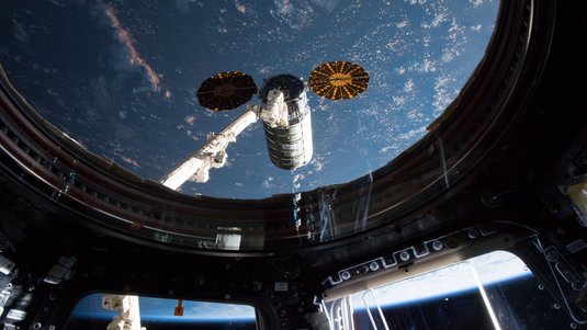 Blick aus dem Inneren der ISS auf ein Modul, das außen befestigt ist; im Hintergrund ist die Erde zu sehen