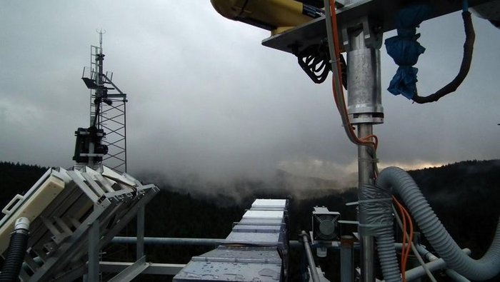 Zu sehen ist ein Teil der Messapparatur, im Hintergrund schiebt sich eine tiefhängende, graue Wolkendecke über einen Bergkamm.