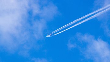 Blauer Himmel, über den ein Flugzeug fliegt, an dessen hinterem Ende sich ein Kondensstreifen zeigt.