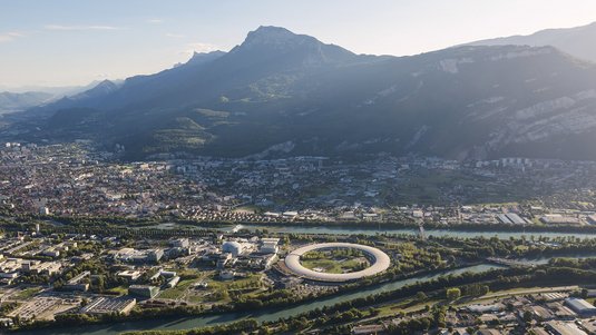 Das Bild zeigt eine Stadt, Berge, einen Fluss und einen großen Beschleunigerring.