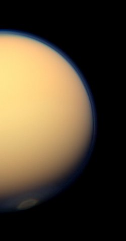 Gelb-orange ragt der Saturnmond als große Halbkugel in das Bild. Die Dunstschicht hebt sich als blaue Hülle um den Mond vor dem schwarzen Hintergrund ab. 