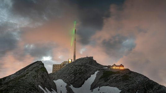 Auf einem Alpengipfel strahlt ein grüner Laser in einen verdunkelten Himmel über einem Antennenmast