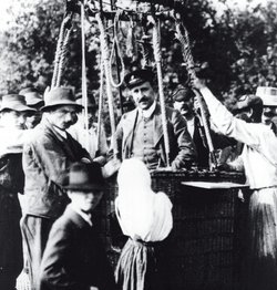 Historisches Foto von Victor Hess kurz vor einem seiner Aufstiege im Forschungsballon. Er befindet sich im Korb von einem Heißluftballon und um ihn herum stehen, außerhalb des Korbes, mehrere Männer und ein Junge.