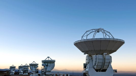 Mehrere Antennen von ALMA stehen vor einer hügeligen Landschaft, während gerade die Sonne aufgeht.