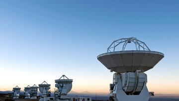 Mehrere Antennen von ALMA stehen vor einer hügeligen Landschaft, während gerade die Sonne aufgeht.
