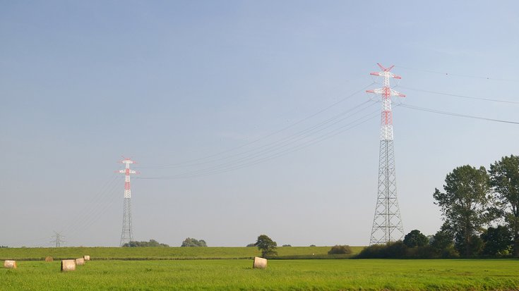 Zwei Strommasten stehen auf einem grünen Feld. Die Hochspannungsleitungen heben sich vor dem blauen Himmel ab.