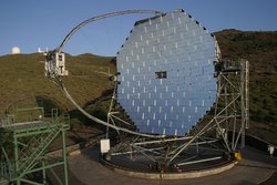 Vor karger Landschaft ein silbriges Gerüst aus einer Ansammlung vieler metallisch glänzender Platten, die gekrümmte Fläche bilden – über diese Fläche wölbt sich ein ovales Gestänge, an dessen Spitze eine Kiste montiert ist.