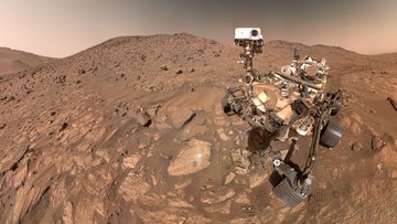 Ein Roboter mit sechs Rädern in einer sandigen, steinigen Wüstenumgebung. In dem Stein vor dem Roboter ist ein winziges Loch. Neben dem Loch ist eine ähnlich große, leuchtend helle Fläche.