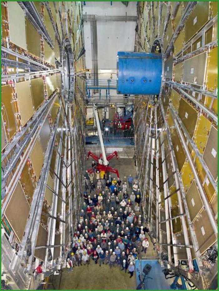 ATLAS-Myonsystem Zwischen zwei 25 Meter hohen Rädern aus plattenförmigen Nachweisgeräten steht eine Gruppe Menschen, die im Verhältnis zur Apparatur auf beiden Seiten winzig wirken. Aus der Mitte des rechten Rads ragt ein blauer Zylinder.