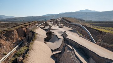 Abilgebildet ist eine Straße, die durch ein Erdbeben zerstört wurde. Die Fahrbahn ist durch längliche Risse zerstört.