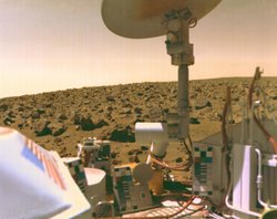 Im Vordergrund sind Teile eines Viking-Landers zu sehen, unter anderem eine Antenne. Im Hintergrund sieht man die mit Geröll übersäte rote Landschaft des Mars mit einem ebenfalls rötlichen Himmel.