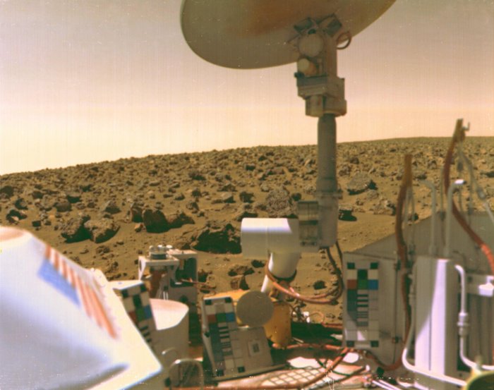 Viking auf dem Mars Im Vordergrund sind Teile eines Viking-Landers zu sehen, unter anderem eine Antenne. Im Hintergrund sieht man die mit Geröll übersäte rote Landschaft des Mars mit einem ebenfalls rötlichen Himmel.
