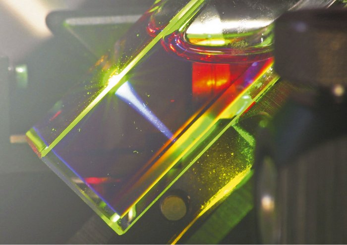 Versuchsaufbau Ein mit einer durchsichtigen Flüssigkeit gefüllter Glasquader, der grünes Licht streut, steht schräg im Bild. An der Seite links oben ein weißer Lichtreflex.