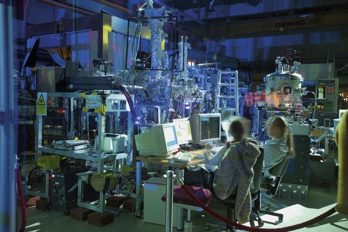Experiment bei FLASH Zwei Wissenschaftler sitzen an einem Schreibtisch, auf dem drei Computermonitore stehen. Hinter den Schreibtisch: viele technische Geräte, teilweise in Alu-Folie eingepackt, sowie eine Leiter, Warnschilder und ein Kunststoffvorhang. Vorne rechts im Bild ein Absperrseil.
