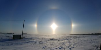 Über einer Eisfläche steht die Sonne tief am Himmel. Um sie herum befindet sich ein Lichtkreis mit zwei hellen Flecken links und rechts von der Sonne. 