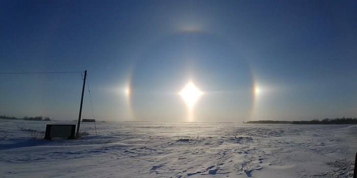 22-Grad-Ring mit Nebensonnen Über einer Eisfläche steht die Sonne tief am Himmel. Um sie herum befindet sich ein Lichtkreis mit zwei hellen Flecken links und rechts von der Sonne.