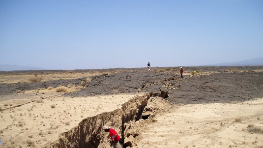 Sandiges Gelände, durch das sich ein etwa ein bis zwei Meter breiter, verkrusteter Riss zieht. Ein Mann mit Hut schaut in den Graben hinunter. 