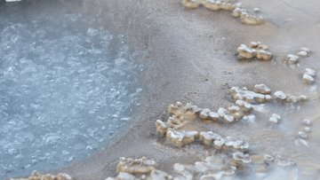 Brodelndes, blau-graues Wasser in einem flachen Becken; am Rand unregelmäßige, weiße Mineralablagerungen auf beige-grauem Boden. Keine Personen, natürliche Umgebung.