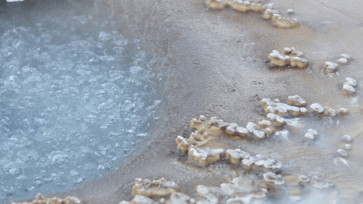 Brodelndes, blau-graues Wasser in einem flachen Becken; am Rand unregelmäßige, weiße Mineralablagerungen auf beige-grauem Boden. Keine Personen, natürliche Umgebung.