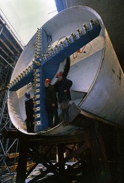 Drei Männer mit Bauhelmen stehen im circa fünf Meter hohen Schneidrad einer zylinderförmigen Tunnelbohrmaschine.