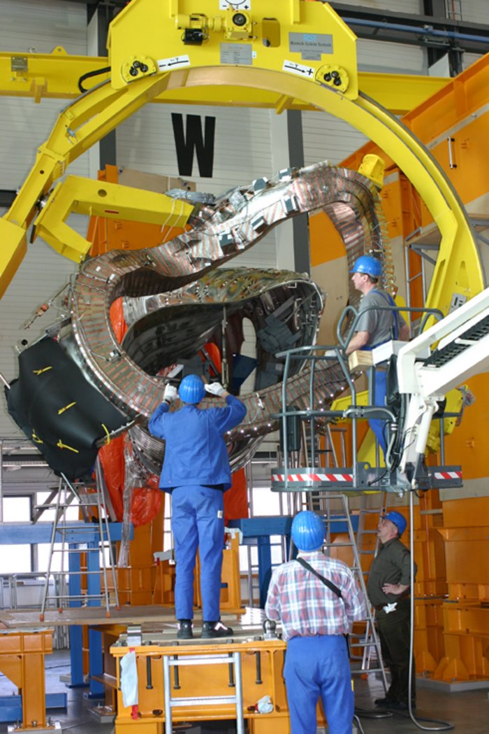 Wendelstein 7-X: Bizarre Magnetspule Das Foto zeigt mehrere Arbeiter, die in einer Monatgehalle eine bizarr geformte Magnetspule aufbauen.