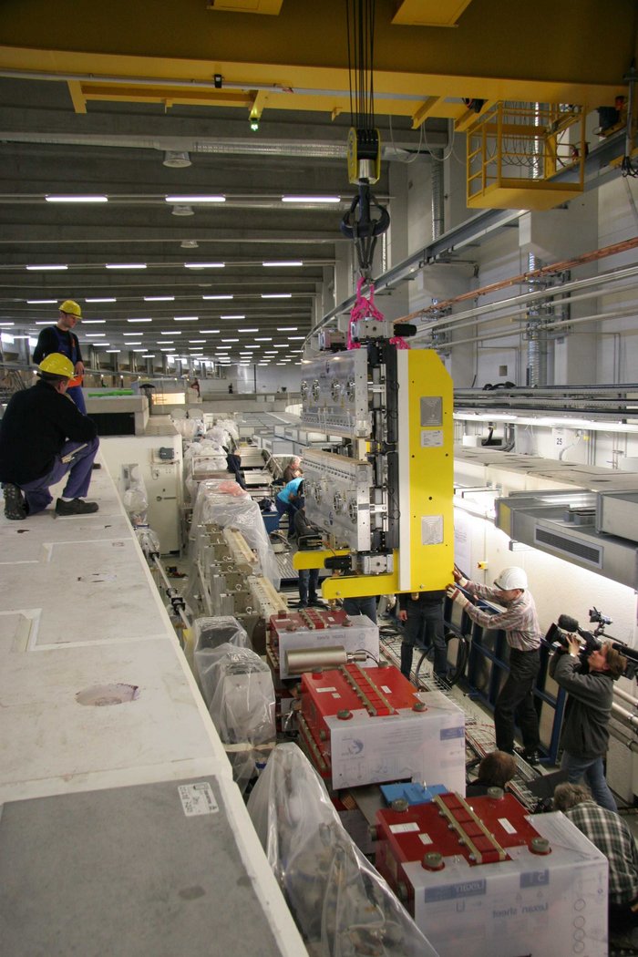 Foto einer schweren Magnetanordnung, die mit Hilfe eines Krans von oben an Ort und Stelle in einem Teilchenbeschleuniger herabgelassen wird. Der Kranführer steht oban auf der Betonabschirmung, die den Beschleuniger umgibt. Mehrere Menschen neben dem Beschleuniger führen die schwere Komponente auf ihrem Weg nach unten.