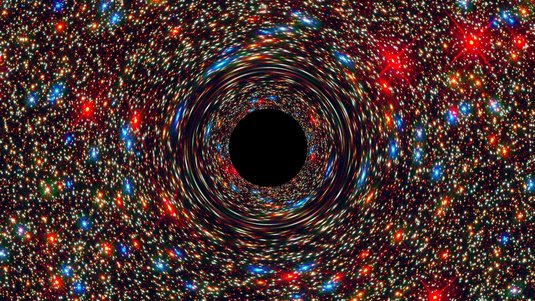 Im Zentrum einer Galaxie mit zahlreichen bunten Sternen und Himmelskörpern befindet sich ein kreisrundes Schwarzes Loch: Die Farbe stellt den Ereignishorizont dar, den keinerlei Materie oder Licht wieder überqueren kann, sobald sie ihn passiert haben.