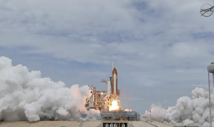 In den ersten Sekunden des Starts hat das Space Shuttle noch kaum von der Startrampe abgehoben. Es hängt an einem großen zigarrenförmigen Treibstofftank, an dessen Seite zwei kleinere, ebenfalls lange zylindrische Feststoffraketen angebracht sind. Aus ihren Enden strömen flammend Antriebsgase. Links ist das Gerüst des Startturms zum Teil zu erkennen. Im Hintergrund steigt der Qualm der Triebwerke hoch.