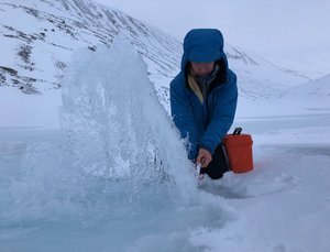 Forscherin Gabrielle Kleber kniet im Schnee eingehüllt in eine blaue Winterjacke hinter einer der Grundwasserquellen, die sie erforscht. Das Wasser sprudelt aus dem Boden und sie entnimmt eine Wasserprobe mit einer Spritze. Im Hintergrund sind schneebedeckte Berge. 