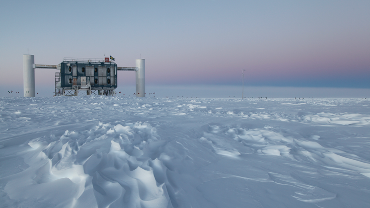 Das Bild zeigt eine Eislandschaft. Im Hintergrund ist die Forschungseinrichtung IceCube zu sehen.