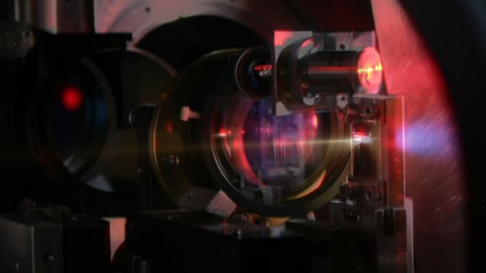 Experiment zur Ionenbeschleunigung Aus schwarz eingefassten runden Linsen tritt das Laserlicht aus. Es trifft auf ein Ziel, an dem ein bläuliches Leuchten sichtbar ist.