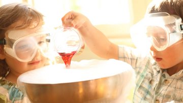 Zwei Kinder mit Schutzbrille füllen eine rote Flüssigkeit in einen Topf