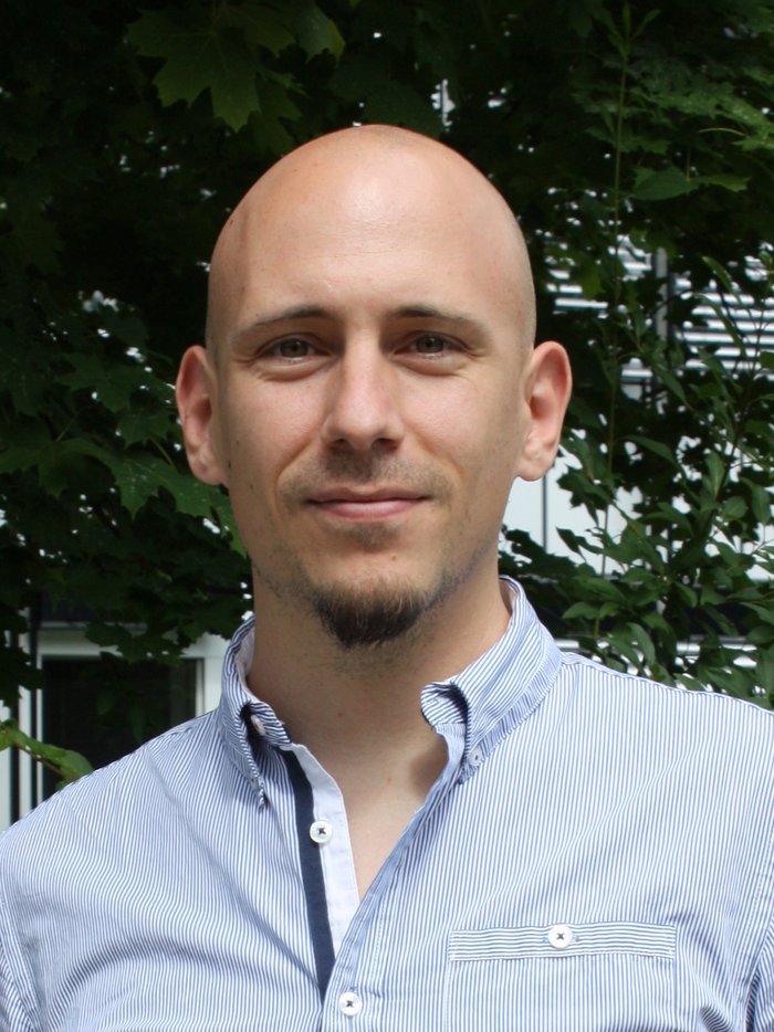 Tobias Wilken vom Max-Planck-Institut für Quantenoptik Ein Mann mit blau-gestreiftem Hemd vor einem grünen Hintergrund aus Blättern.
