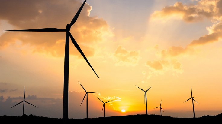 Foto mehrerer Windräder in hügeliger Landschaft bei tiefstehender Sonne.