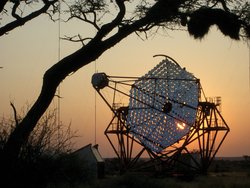 Das Bild zeigt eines der H.E.S.S.-Teleskope, dessen Spiegelfläche aus zahlreichen kleineren, runden Spiegeln zusammengesetzt ist. Im Hintergrund ist der Sonnenuntergang in einer Steppenlandschaft zu sehen.