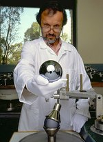 In einem Labor hält ein Mann im weißen Kittel und mit weißen Stoffhandschuhen hält eine metallisch-spiegelnde Siliziumkugel von etwa zehn Zentimetern Durchmesser in der Hand.