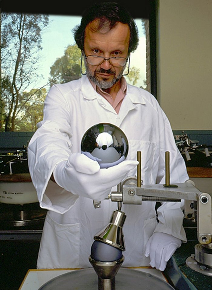 In einem Labor hält ein Mann im weißen Kittel und mit weißen Stoffhandschuhen hält eine metallisch-spiegelnde Siliziumkugel von etwa zehn Zentimetern Durchmesser in der Hand.