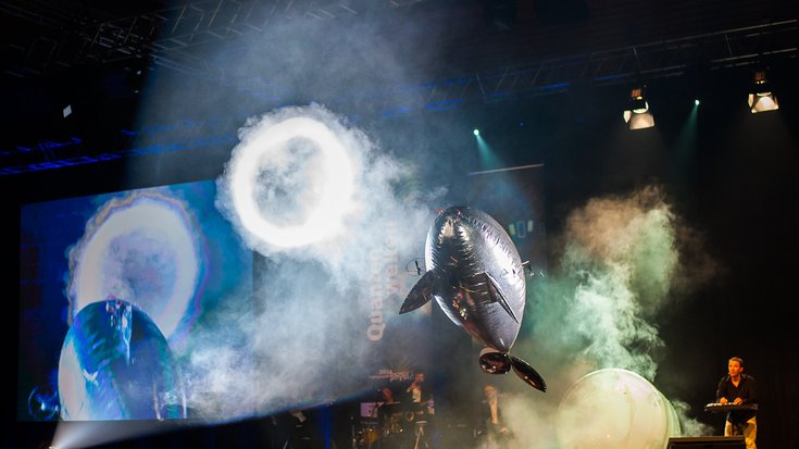 Ein Publikum sitzt in einem großen Saal und von der Bühne aus sind Ringe aus Nebel und ein fliegender Ballon zu sehen.