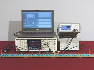 Technisches System: Laptop steht auf einer Apparatur mit vielen Schaltern und einem kleinen Display; rechts daneben ein weiteres Gerät mit Anzeige; davor eine Steckerleiste mit verschiedenen Kabeln